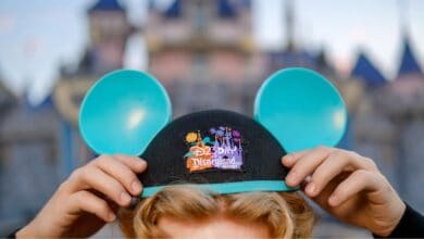 Blue Mickey ears hat featuring the iconic Disneyland logo held up proudly, with Sleeping Beauty Castle softly blurred in the background—an exclusive D23 moment capturing pure Disney magic and fan excitement.