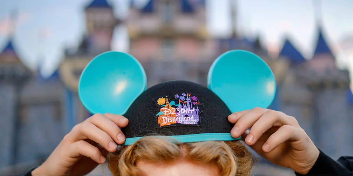 Blue Mickey ears hat featuring the iconic Disneyland logo held up proudly, with Sleeping Beauty Castle softly blurred in the background—an exclusive D23 moment capturing pure Disney magic and fan excitement.