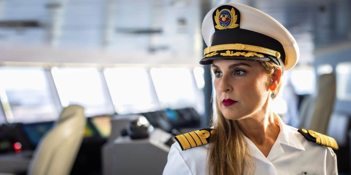 A Disney Cruise Line captain in crisp white uniform and hat stands confidently on the ship’s bridge, gazing out as she prepares for another magical voyage at sea.