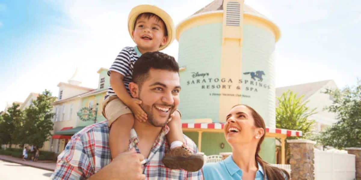 Happy family outside Disney’s Saratoga Springs Resort, child riding high on dad’s shoulders while mom gazes up with a big smile, all enjoying exclusive Disney Vacation Club member perks like complimentary snacks during their magical July 22 Disney getaway.