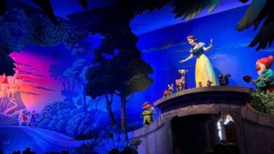 Snow White greets visitors from a picturesque bridge in Fantasyland, surrounded by friendly forest animals and one of the Seven Dwarfs, with the fairytale castle and glowing sunset completing the magical scene.