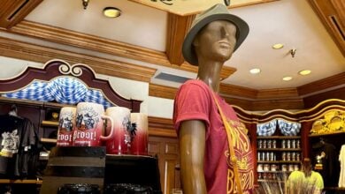 A mannequin dressed in a festive red shirt and hat welcomes guests near rows of souvenir mugs inside the bright, cheerful gift shop at EPCOT's Germany Pavilion.