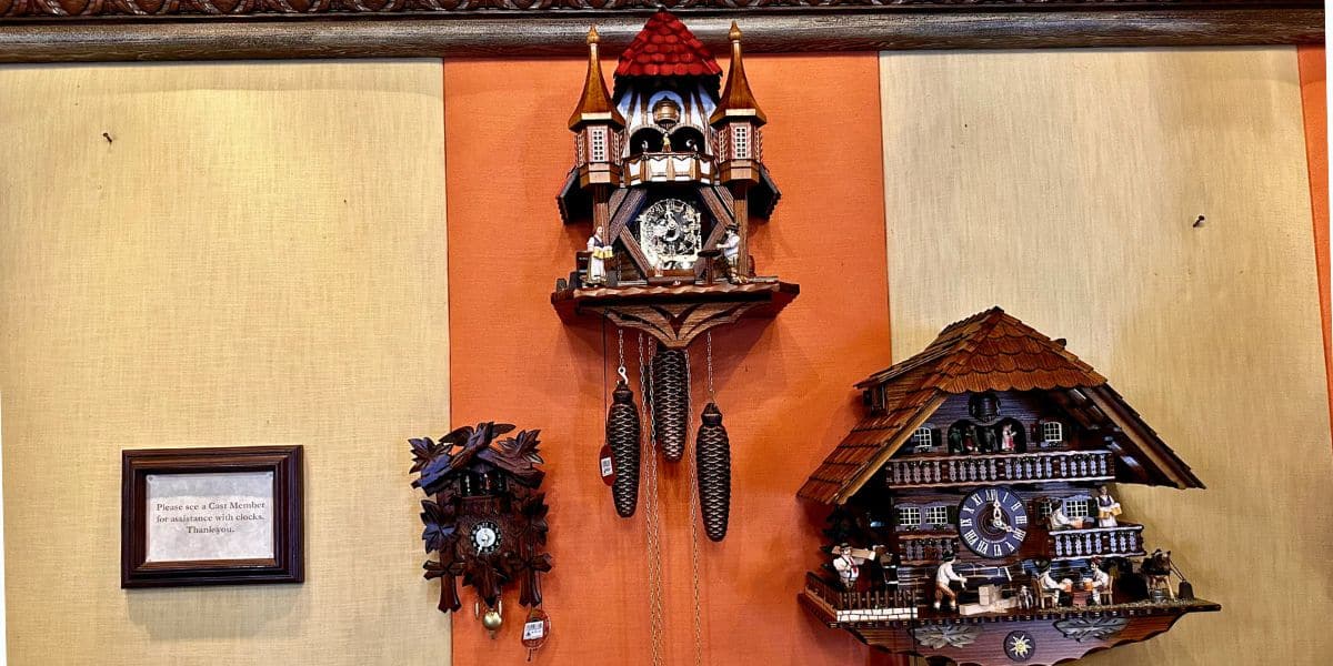 Three whimsical cuckoo clocks, just like the ones found in the charming shops of EPCOT’s Germany Pavilion, decorate a striped orange and beige wall above a framed sign—capturing that quintessential World Showcase magic.