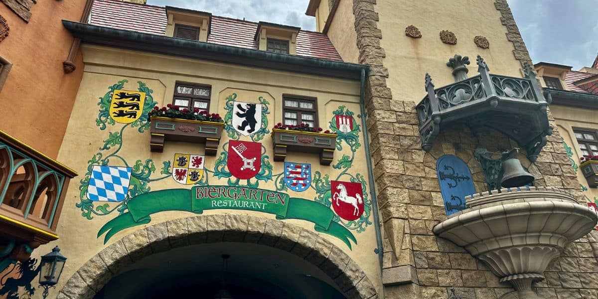 The vibrant facade of the Biergarten Restaurant in EPCOT’s Germany Pavilion features ornate crests, a balcony overlooking the plaza, and a whimsical decorative fountain, welcoming guests into an immersive Oktoberfest celebration.