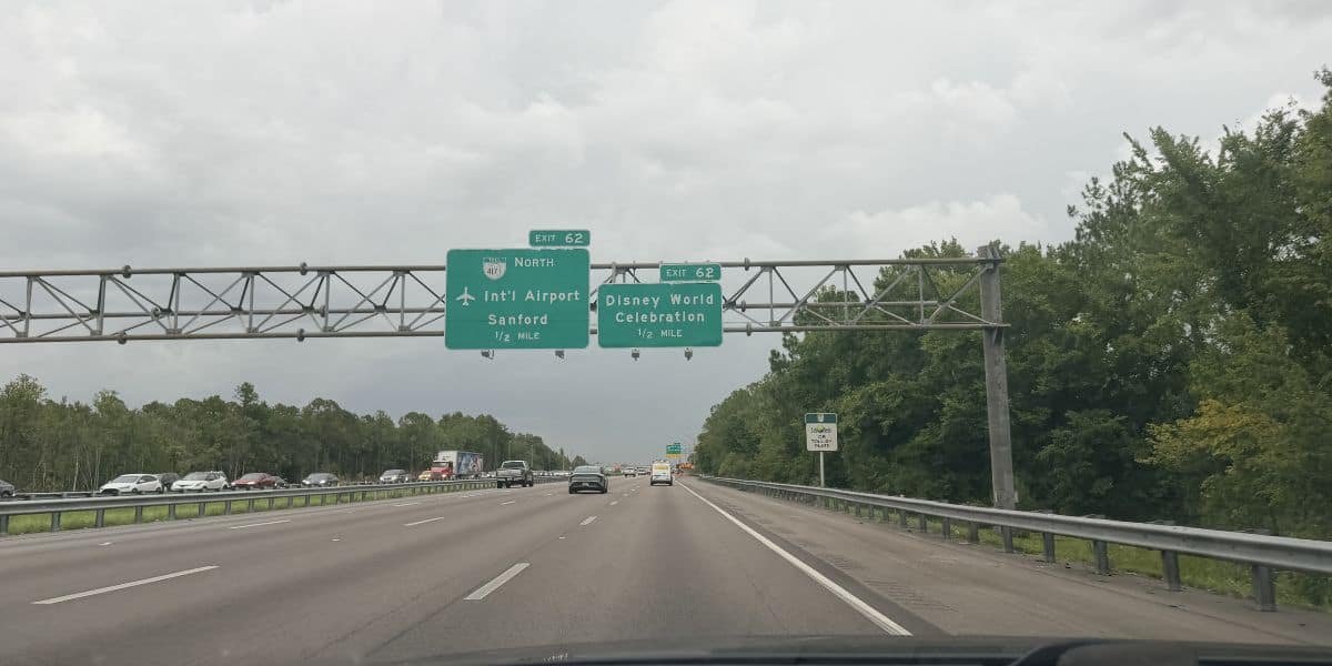 Highway leading to Disney World, with green exit signs pointing the way for excited visitors; a Tesla and other cars drive toward the magic, flanked by lush trees on both sides.