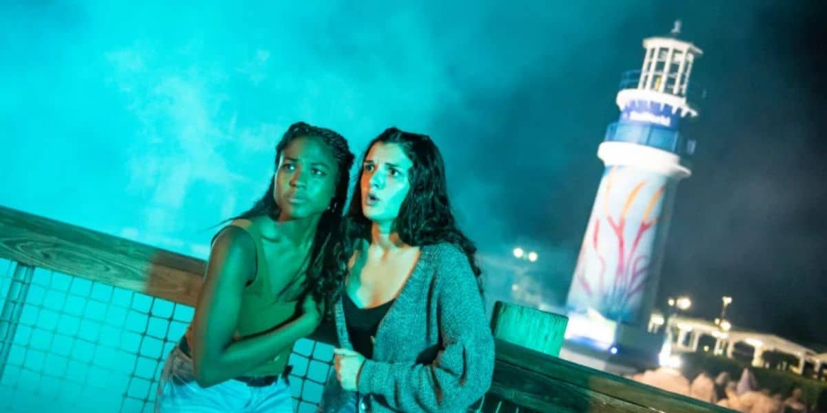 Guests huddle in fear by a fence under the eerie glow of SeaWorld Orlando’s Halloween Horror Nights, with the iconic lighthouse illuminated against a backdrop of swirling fog and blue light, heightening the chilling thrills of this spine-tingling event.