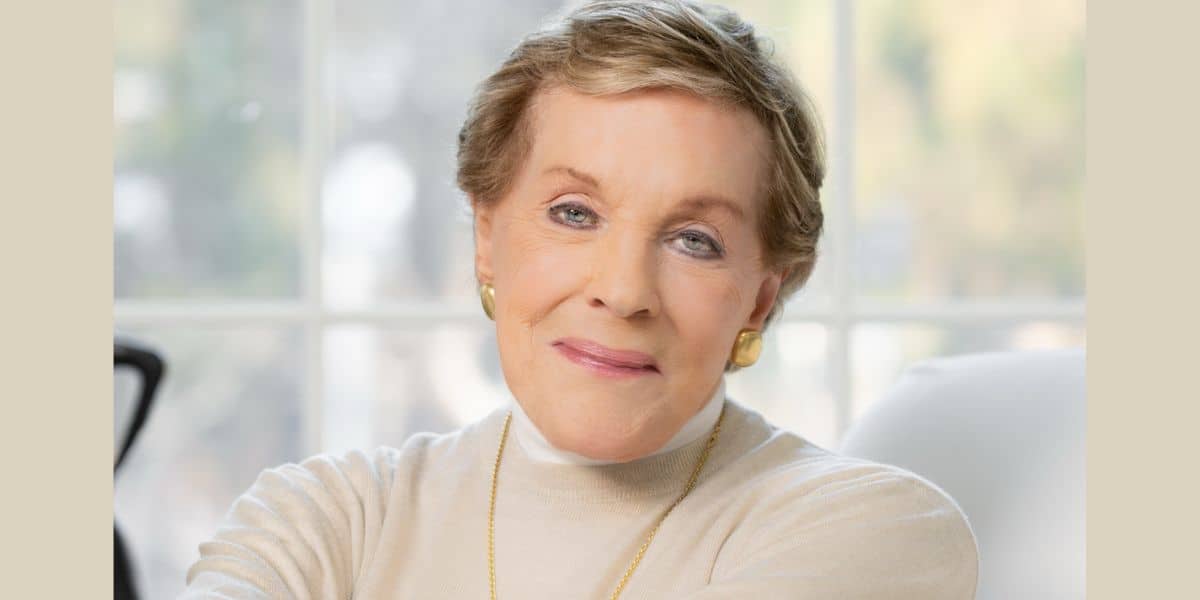 A cheerful older woman with short light hair and a cream sweater beams warmly inside a sunlit theme park lounge, exuding the welcoming spirit of your favorite resort host.