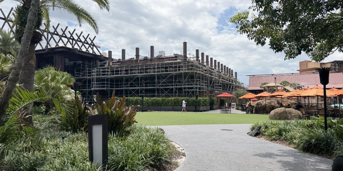A section of the theme park is being built, with scaffolding surrounding the new attraction. Nearby, palm trees sway and guests relax at brightly colored tables shaded by orange umbrellas.