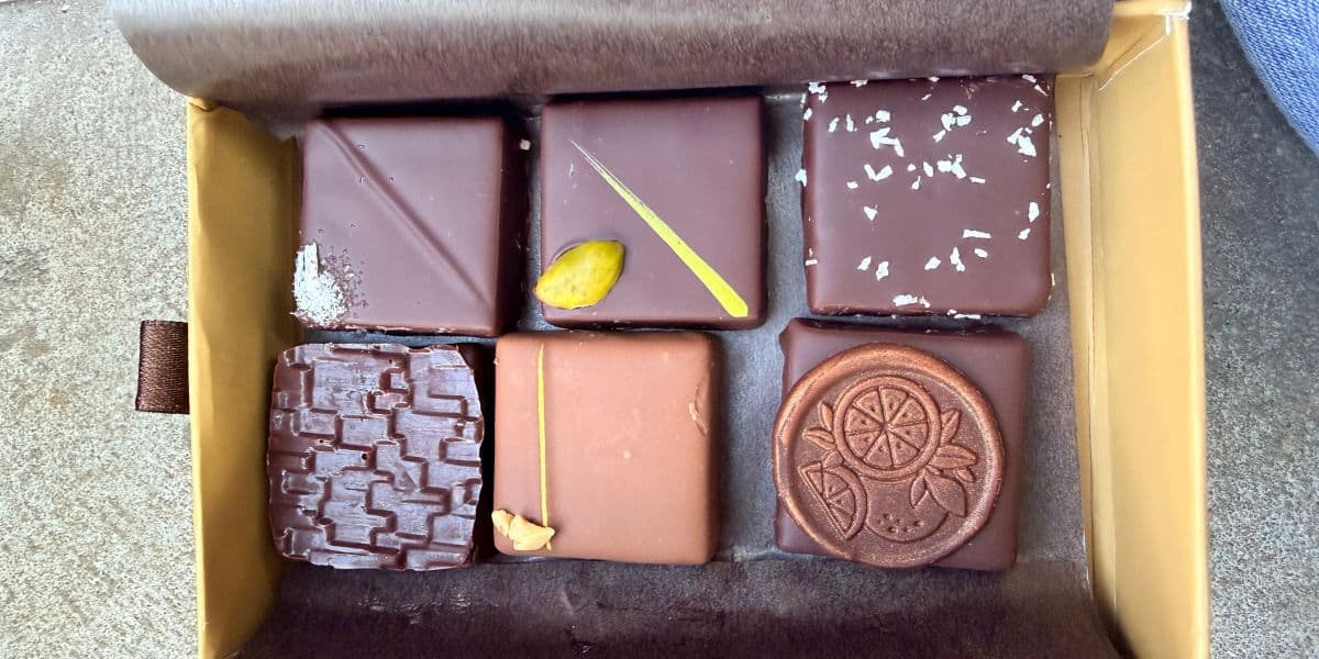 A selection of seven artisan chocolates, each decorated with whimsical Disney-inspired toppings and designs, displayed in a festive box on a brown and tan backdrop—an ideal treat to enjoy during Halloween festivities or to relive the magic of Disney Springs at home from your visit to Walt Disney World.