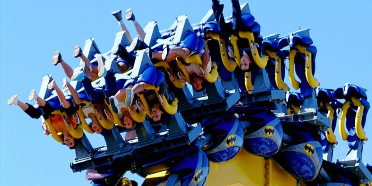 Thrill-seekers soar on the inverted Batman: The Ride roller coaster at Six Flags, legs flying free beneath them against a brilliant blue sky.