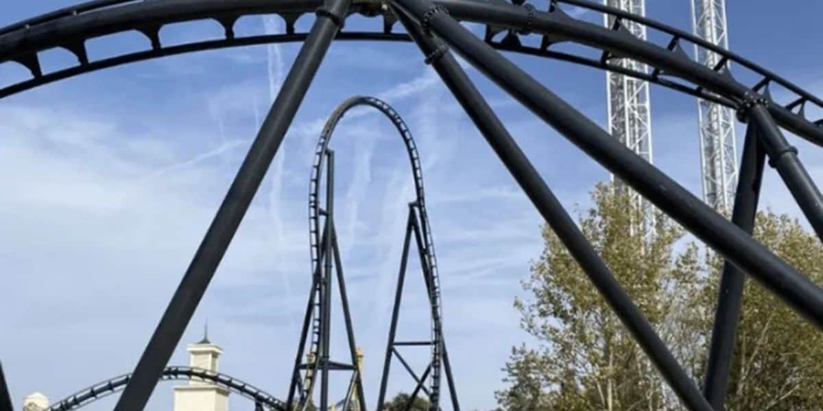Soaring black steel coaster tracks and thrilling loops curve against a bright blue sky, with lush trees lining the ride at the amusement park.