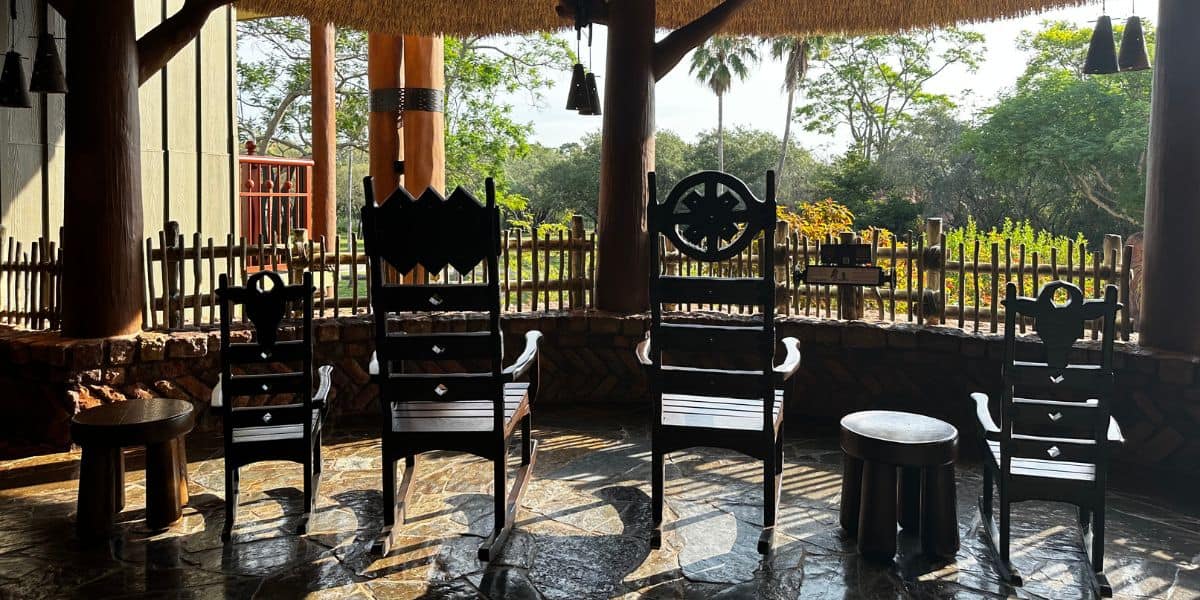 Four ornate wooden chairs and two round stools await in a sun-drenched, rustic outdoor lounge surrounded by leafy trees, channeling the deluxe resort vibes of Disney World’s most relaxing amenities—an inviting retreat for guests seeking magical downtime during an October 2026 visit.