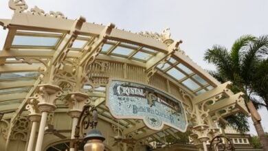 The grand entrance of Magic Kingdom’s Crystal Palace dazzles with its ornate Victorian trim and sparkling glass roof, while palm trees sway in the enchanting backdrop, inviting guests into a whimsical world of dining and Disney magic.