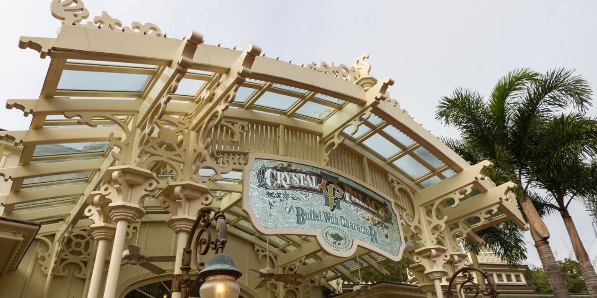 The grand entrance of Magic Kingdom’s Crystal Palace dazzles with its ornate Victorian trim and sparkling glass roof, while palm trees sway in the enchanting backdrop, inviting guests into a whimsical world of dining and Disney magic.