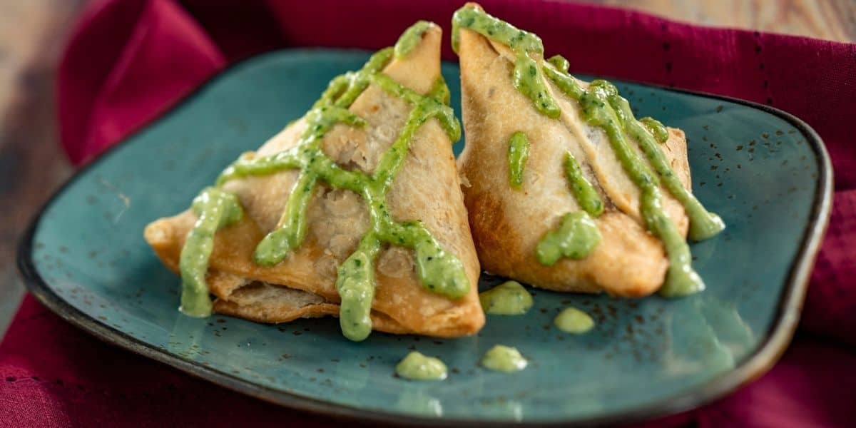 Two golden-brown samosas served on a vibrant blue plate, drizzled with green chutney, ready for theme park visitors to enjoy against a plush burgundy backdrop.