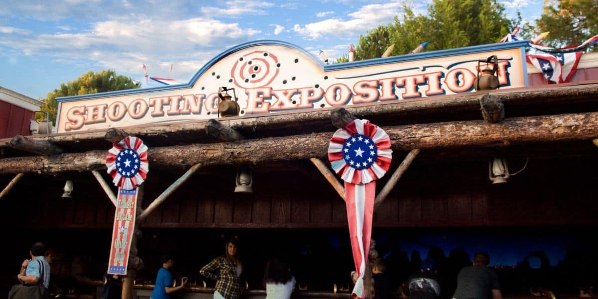 Frontierland Shooting Exposition sign adorned with red, white, and blue decorations as Guests gather beneath the themed wooden canopy.