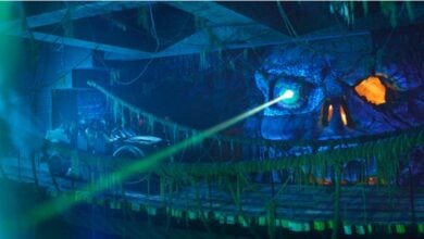 An Indiana Jones Adventure ride vehicle races past a glowing skull cave radiating eerie green light, while dynamic blue and orange lighting amps up the excitement—showcasing new thrills as this iconic Disneyland attraction returns with electrifying updates.