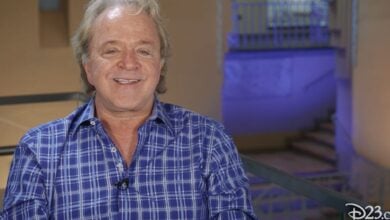 Smiling voice actor in a blue plaid shirt sits inside, with illuminated stairs behind him—he’s well known among Disney fans and frequently celebrated as a Disney Legend.