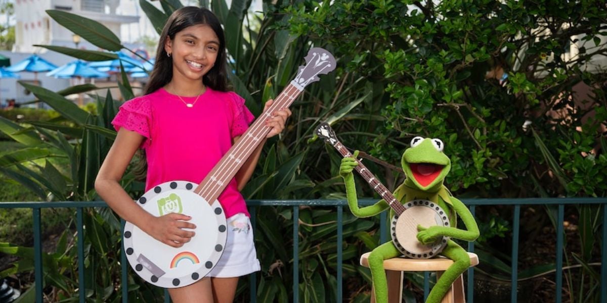 A young visitor with a toy banjo poses beside Kermit the Frog, who strums his own banjo in an outdoor setting at a family entertainment park.