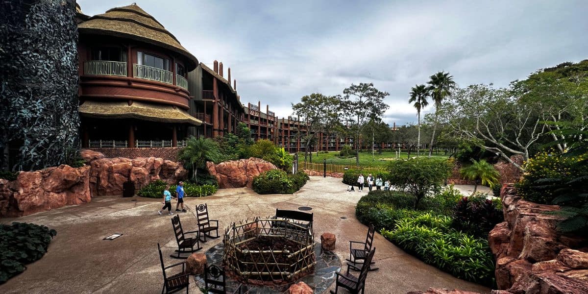 Guests stroll past a glowing fire pit encircled by rocking chairs, nestled among vibrant greenery and impressive Deluxe Resort buildings at Disney World. This photo captures the relaxing ambiance and upscale amenities awaiting visitors during their October 2026 getaway.
