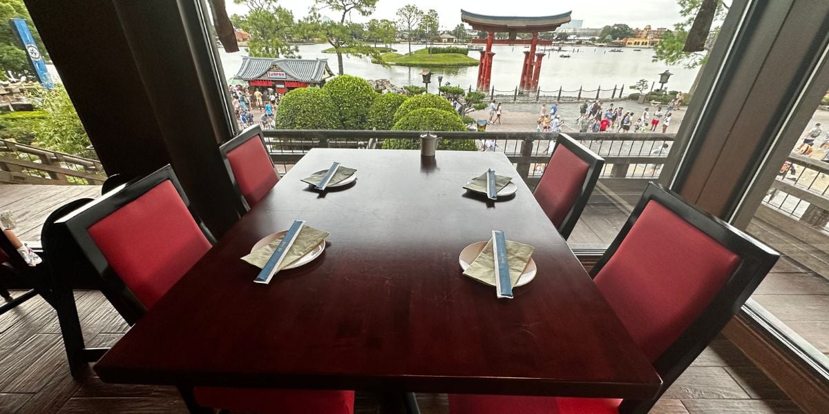 Dining table with four red chairs set inside a Japan Pavilion-inspired restaurant at EPCOT, overlooking the World Showcase lagoon and the iconic large red torii gate through expansive windows—offering the perfect immersive atmosphere for theme park fans seeking an upgraded Disney dining experience.