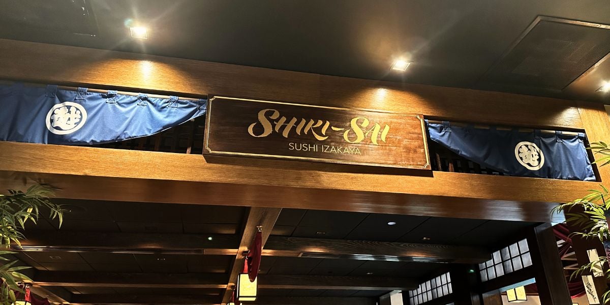 A wooden sign with “SHIRI-SHI SUSHI IZAKAYA” hangs above blue curtains inside a Japanese-inspired restaurant, capturing the immersive, authentic atmosphere you’d expect in Disney World’s EPCOT Pavilion—perfect for theme park fans seeking a new culinary adventure reminiscent of their favorite entertainment destinations.