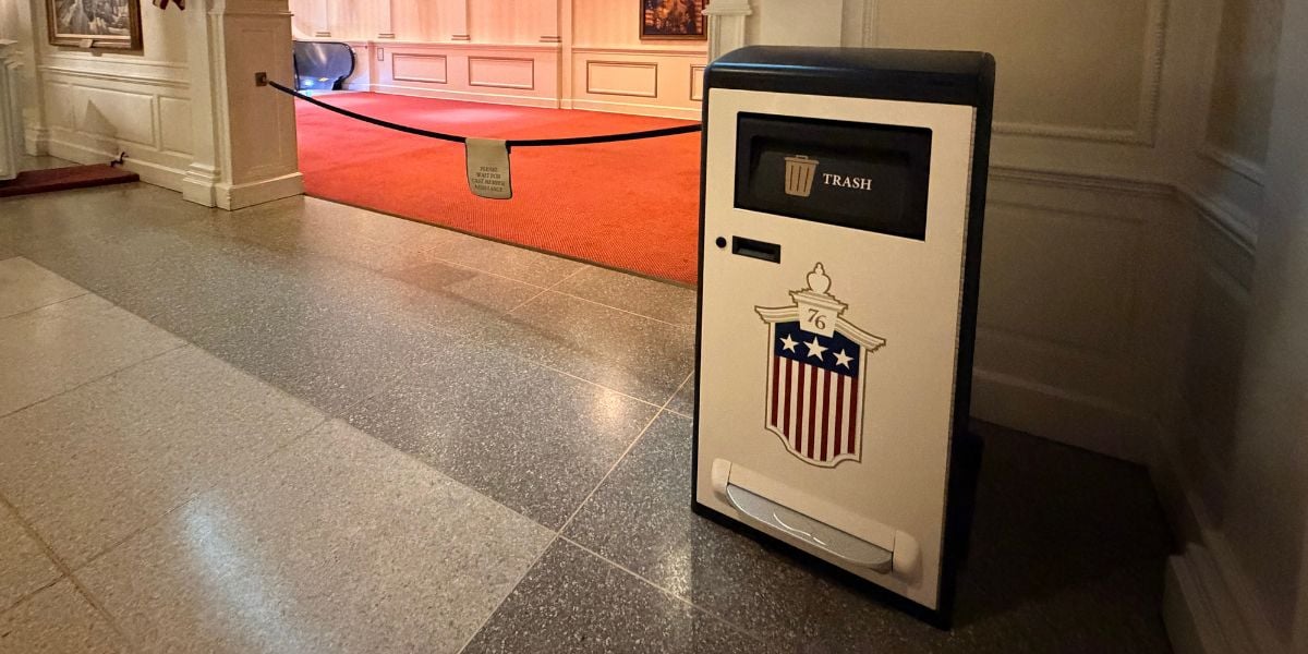 Classic Disney theming shines with this white trash can featuring a patriotic red, white, and blue shield design, perfectly at home in a hallway lined with red carpet and Disney artwork—a playful nod to the whimsical Trash Can Takeover décor at Animal Kingdom.