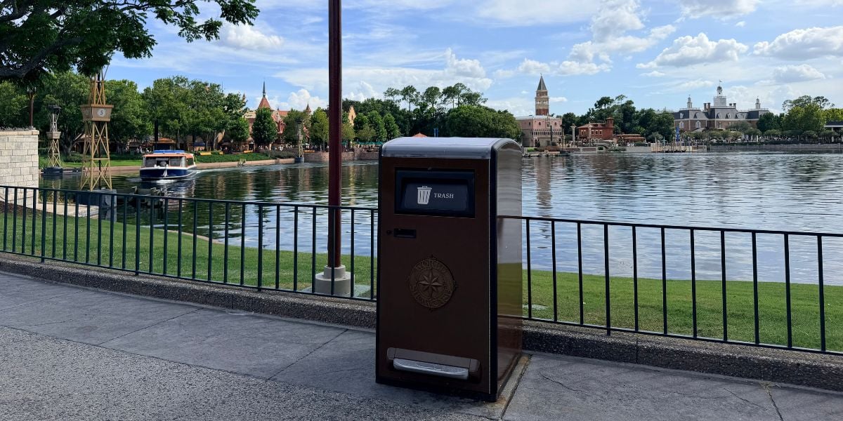 Classic Walt Disney World brown trash can along a scenic lakeside walkway, surrounded by iconic resort buildings and lush trees under a picturesque partly cloudy sky.