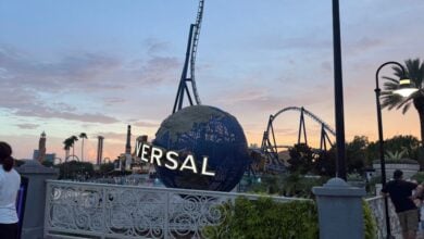 The iconic Universal globe surrounded by thrilling roller coasters and swaying palm trees at sunset, with guests strolling through Universal Studios’ lively entrance area.