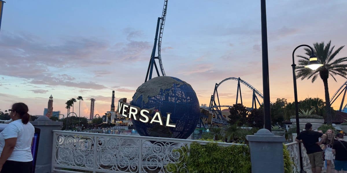 The iconic Universal globe surrounded by thrilling roller coasters and swaying palm trees at sunset, with guests strolling through Universal Studios’ lively entrance area.