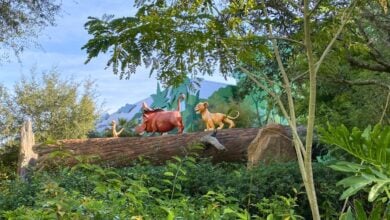 Pumbaa and Simba statues atop a log immerse guests in Disney’s Art of Animation Resort, surrounded by vibrant greenery and trees beneath a bright blue sky.