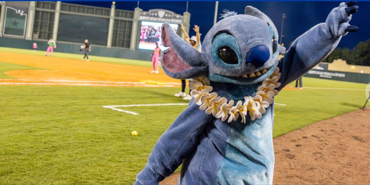 Stitch entertains fans on the baseball field at the 2026 Banana Bowl Championship, cheerfully waving and sporting a vibrant flower lei, adding a splash of Disney+ magic to the big game atmosphere.