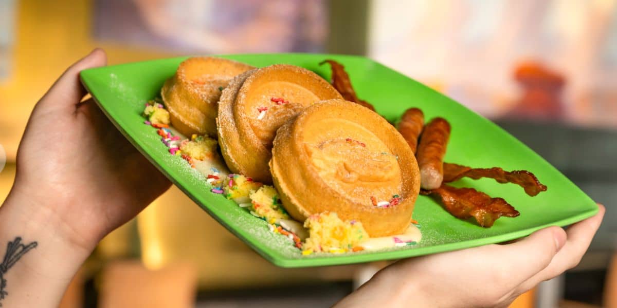 A colorful stack of sprinkle-topped pancakes alongside bacon strips and sausages on a vibrant green plate, ready to enjoy as part of a fun-filled day at your favorite theme park eatery. Two hands excitedly hold the plate, capturing the playful energy of a theme park breakfast.