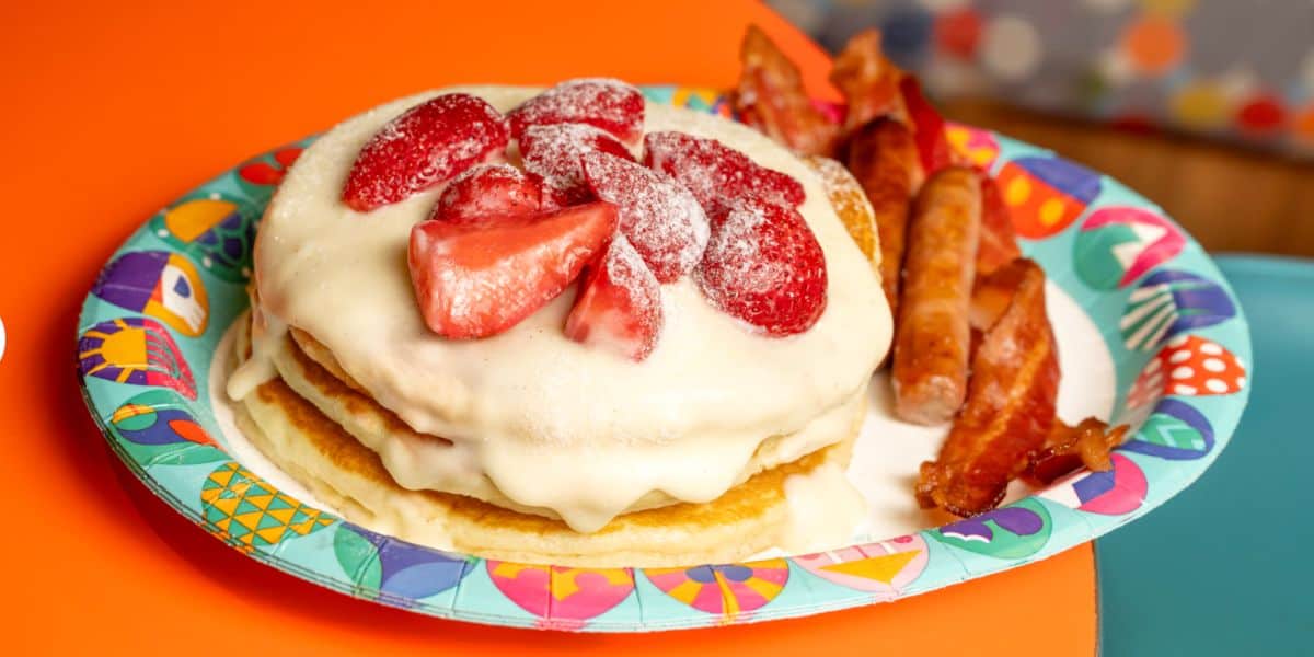 A festive stack of pancakes topped with white icing and fresh strawberries, paired with bacon and sausage, served on a vibrant plate that looks straight out of a character breakfast at Disney World.