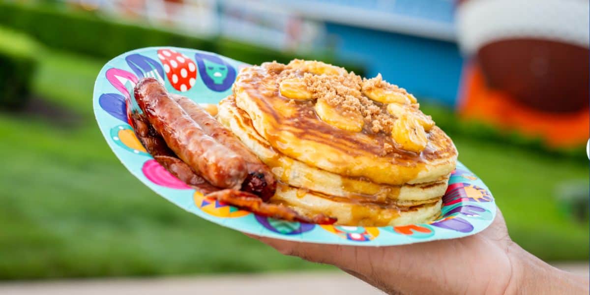 A guest enjoys a vibrant plate loaded with fluffy pancakes, banana slices, golden syrup, sweet crumbles, and two sizzling sausages at a bustling theme park café.