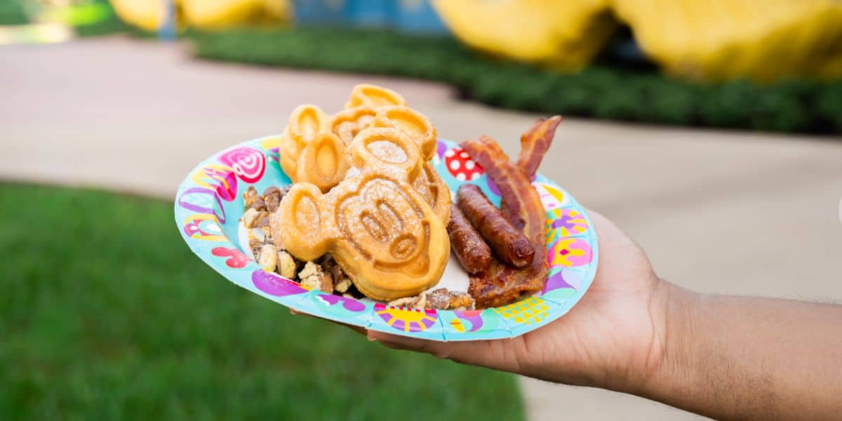 A plate of classic Disney breakfast favorites—Mickey Mouse-shaped waffle, crispy bacon, savory sausage, and fluffy scrambled eggs—is ready to enjoy outdoors at Disney World.