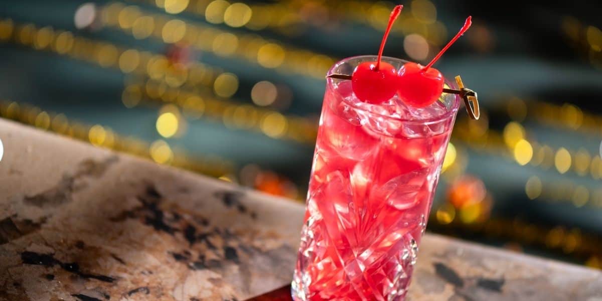A vibrant pink cocktail with ice and two cherries on top, served in a decorative glass on marble—channeling the festive, whimsical vibe of the Hurly-Burly at Disney World’s lively BoardWalk Lounge.