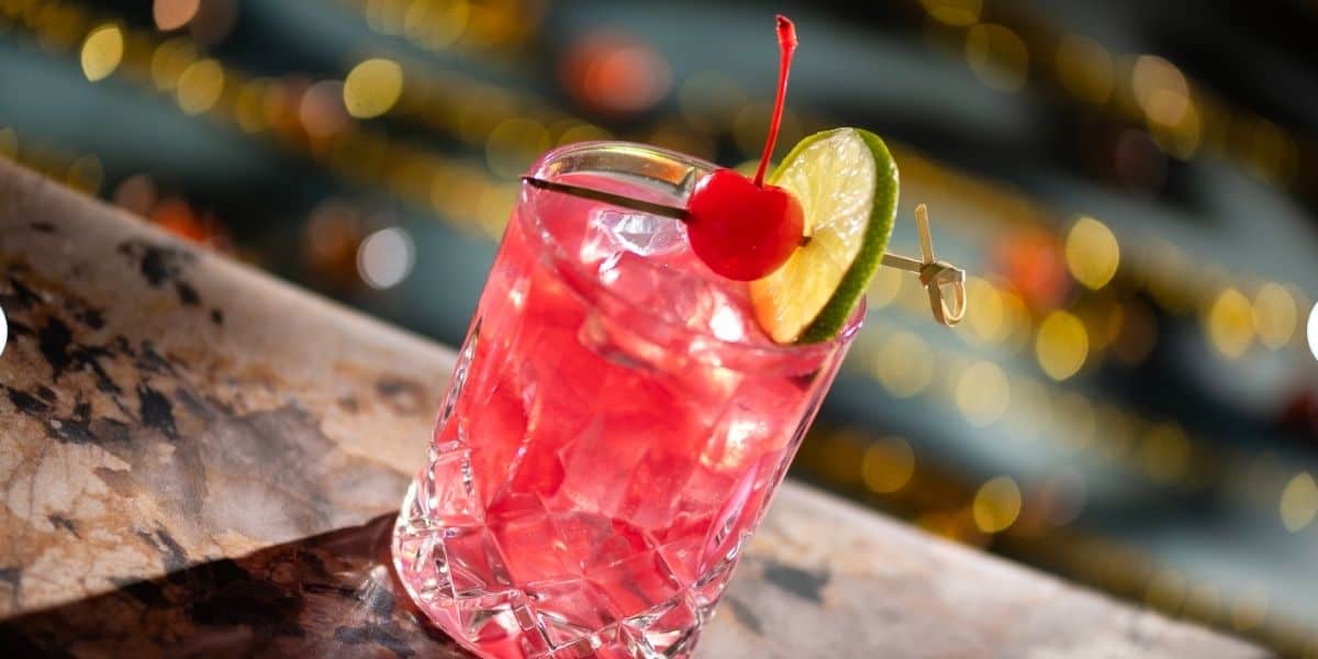 A vibrant pink cocktail with ice, topped with a cherry and lime slice, sits on a sleek marble counter surrounded by twinkling lights—an ideal refresher to sip at BoardWalk Lounge in Disney World after riding your favorite attractions all day.
