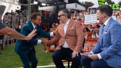 ESPN hosts shake hands on the College GameDay set, surrounded by cheering fans holding signs and another host in the background.