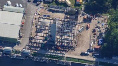 Aerial view of a theme park under construction with steel ride frameworks, colorful machinery assembling attractions, and existing entertainment venues nearby.