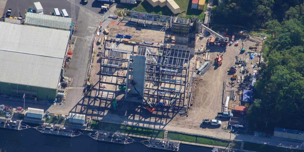 Aerial view of a theme park under construction with steel ride frameworks, colorful machinery assembling attractions, and existing entertainment venues nearby.
