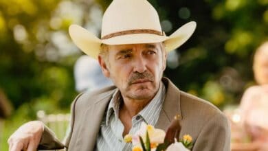 A sharp-dressed cowboy character in a white hat and suit jacket sits outside in a lush, themed park setting, smiling with a bouquet of flowers—evoking the classic charm and relaxed hospitality of the Wild West experience.