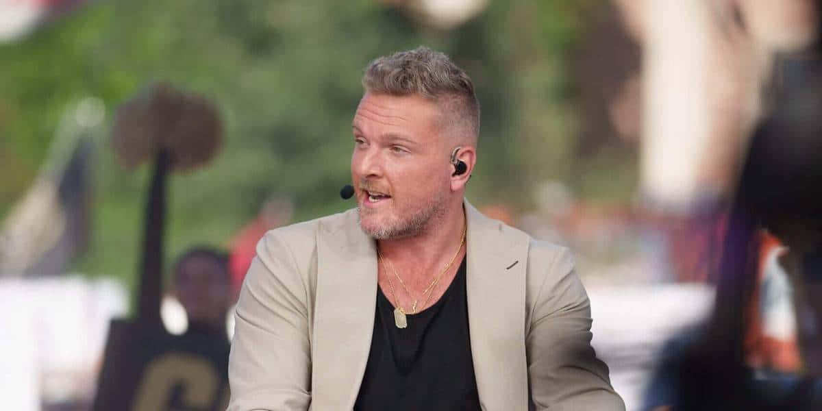 Theme park host with short blonde hair and beard, sporting a headset, beige jacket, and black shirt, speaks with guests outside, sharing his take on double standards in sports coverage—a hot topic often brought up by personalities like Pat McAfee.