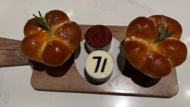 A delicious preview of Steakhouse 71 at Disney’s Contemporary Resort: two round rosemary-topped bread rolls and two mini desserts—one featuring the “71” logo—beautifully presented on a wooden board. Reservations are a must for this popular Walt Disney World dining spot.