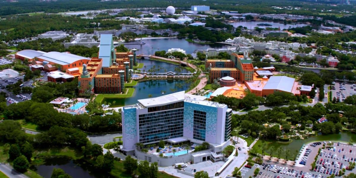 Aerial view of the Walt Disney World resort complex featuring multiple themed hotels, sparkling pools, and a lushly landscaped lake, with visible progress on the new convention center expansion adjacent to one of Disney’s signature resort hotels.
