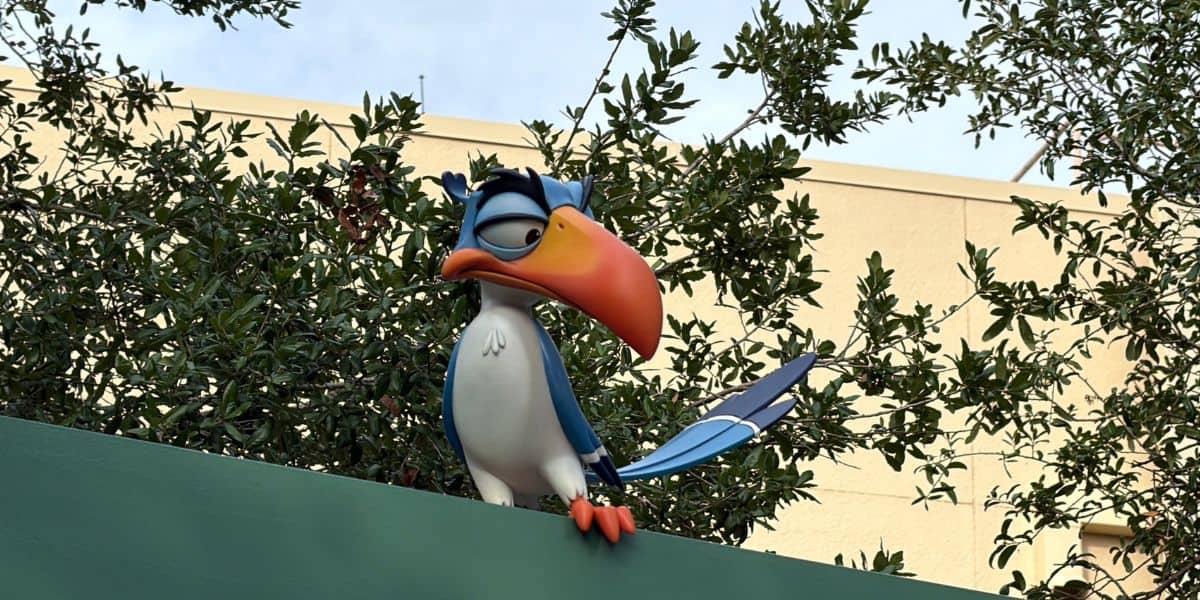 A whimsical cartoon bird with a vibrant orange beak perches on a bright green ledge, framed by lush leafy branches, capturing the playful energy and nostalgic appeal of beloved Disney park characters.