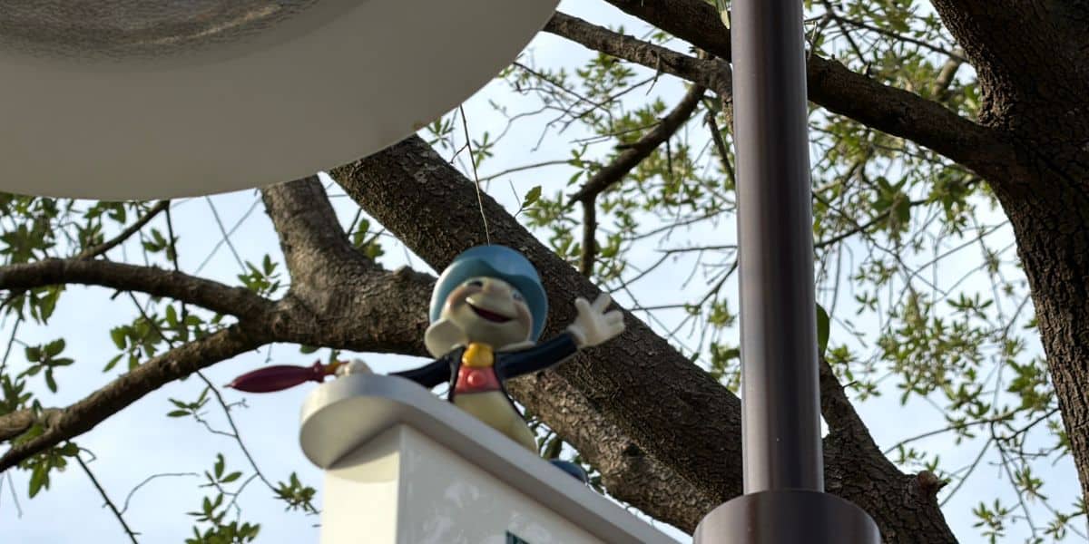 Jiminy Cricket, sporting his signature blue hat, greets guests with a cheerful wave from atop a white sign surrounded by lush greenery and a classic lamppost.