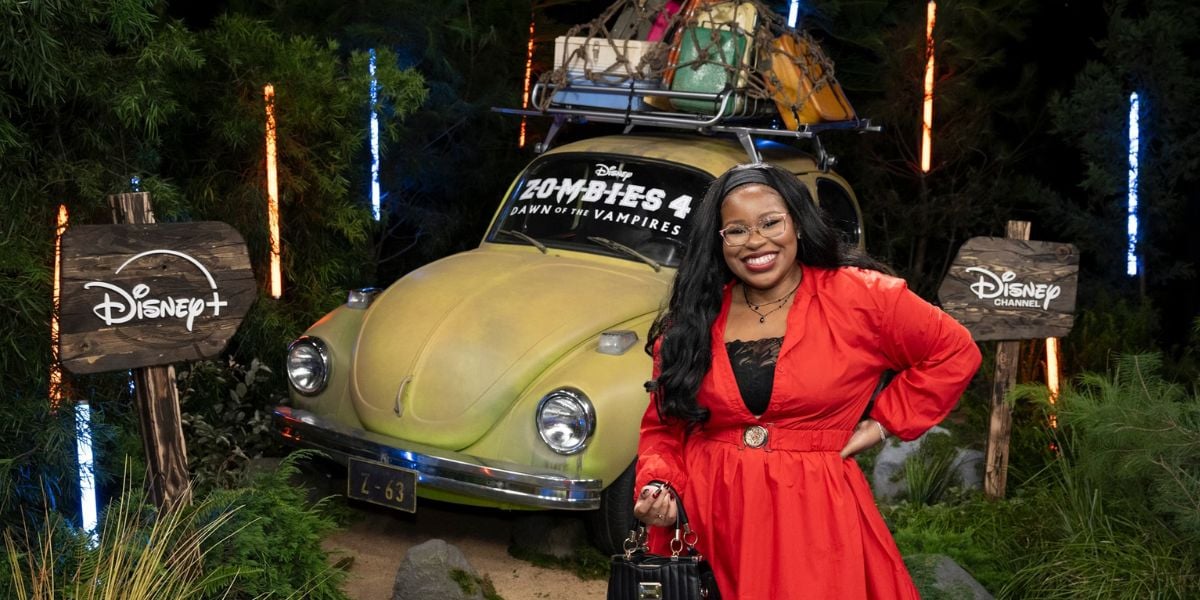 Bree from Disney Zombies strikes a pose in a vibrant red dress beside a yellow car and colorful “Zombies 4” signage, set amid eye-catching Disney+ and Disney Channel displays—capturing the fun, immersive energy of a theme park entertainment showcase.