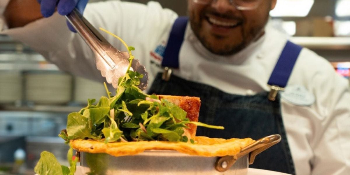 A Chef de Cuisine uses metal tongs to place fresh greens onto a dish at a Disney World restaurant.