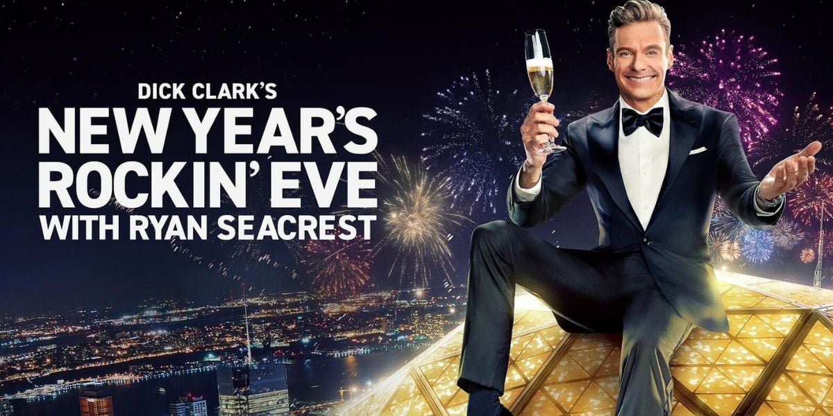 Ryan Seacrest sits on the New Year’s Rockin’ Eve ball, holding a branded champagne glass amidst the fireworks display.